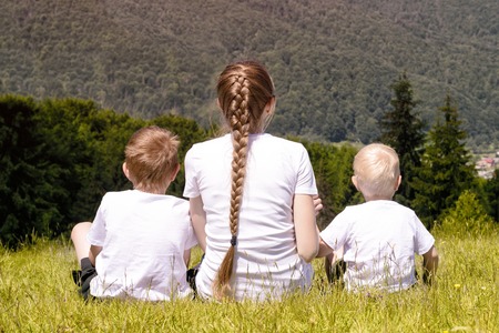 Mother and two young sons sitting on grass on the background of coniferous forest and mountains.の写真素材