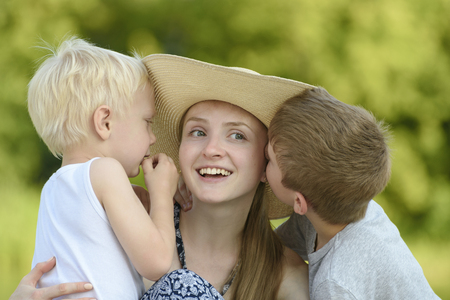 Two little sons hug the mother and kiss on the cheek against the backdrop of greenery.の写真素材