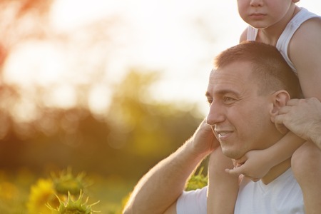 Happy father with his son on their shoulders walking along a green field of blooming sunflowers at sunset. Close-up.の写真素材