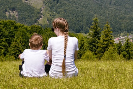 Mother and young son sitting on grass on the background of coniferous forest and mountains.の写真素材