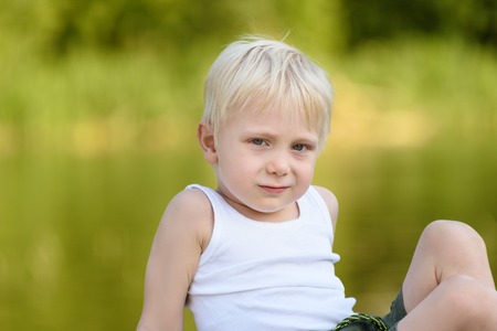Little blond boy sitting outdoors on the background of blurry green leavesの写真素材