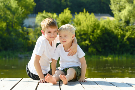Two little boys sit in an embrace on the banks of the river. Concept of friendship and fraternity.の写真素材
