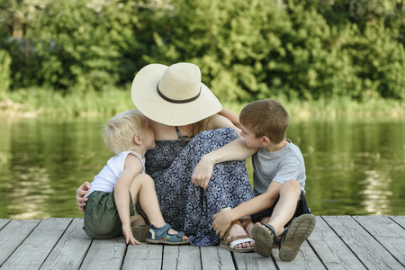 Two little son kissing and hugging her mother sitting on a pier at the river bank.の写真素材