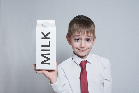 little blond boy holds and shows a big white carton milk package. White shirt and red tie. Light background.の写真素材