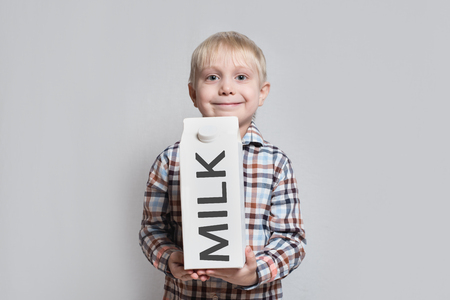 Happy little blond boy is holding a big white carton milk package. Light backgroundの写真素材