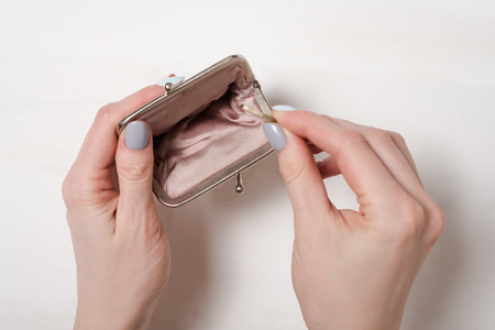 Hand puts a coin in an empty open metal purse on a light background.