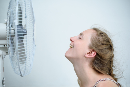 Young woman sitting under a fan with closed eyes from pleasure. Summer heat. White backgroundの写真素材