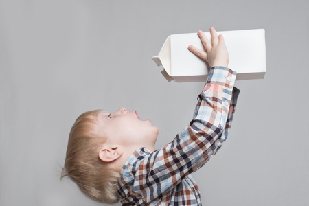 Little blond boy drinks from a large white package. Light background.の写真素材