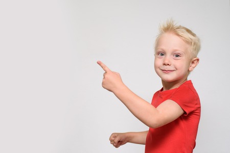 Little blonde boy in red t-shirt points his finger. Space for text. Place for advertising. White backgroundの写真素材