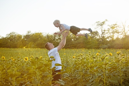 Father throws up his little son on sunny sunflowers field.