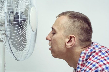 Man with stubble on his face suffers from the heat and trying to cool off near the fan.の写真素材