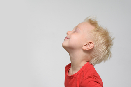 Little blond boy in a red T-shirt with closed eyes from pleasure.. Space for text. White backgroundの写真素材