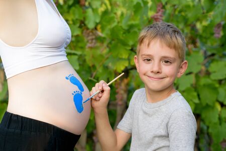 Happy little boy draws with a brush on the belly of his pregnant mom baby's footprint. Pregnant concept. Close-upの写真素材