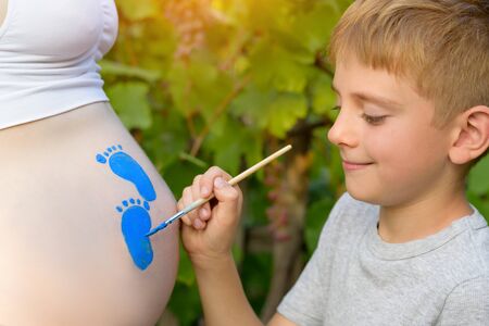 Little boy draws with a brush on the belly of his pregnant mom baby's footprint. Pregnant concept. Close-upの写真素材