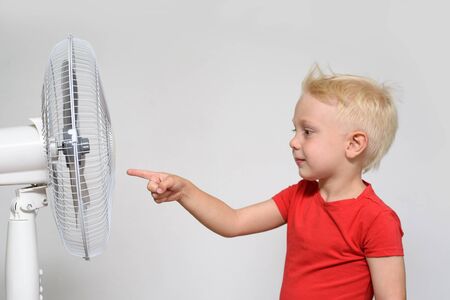 Little blond boy in a red t-shirt touches the fan with his finger. Summer conceptの写真素材