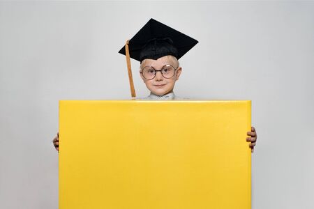 Blond boy in an academic hat and glasses holds a large box. White background. School conceptの写真素材