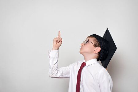 Boy in a square academic hat and glasses points his finger up. School concept. White background.の写真素材