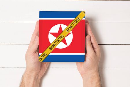 Yellow tape with the inscription Sanctions on the box with the flag of Noth Korea in male hands. Top...
