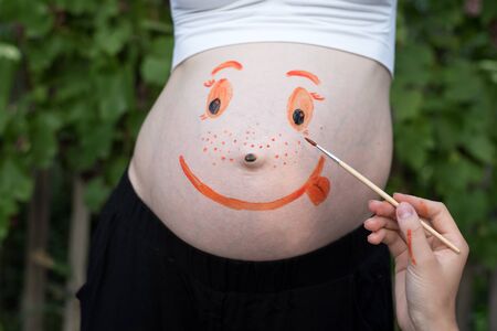 Hand with a brush paints on the belly of a pregnant woman's smiling face. Green backgroundの写真素材
