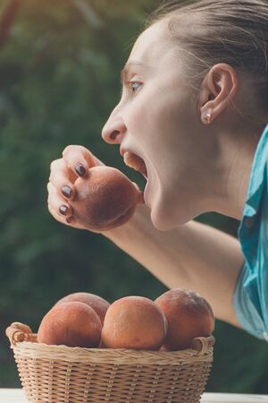 Young woman bites a ripe peach. Wicker basket, green garden on the background. Close-up. Fruit seasonの写真素材