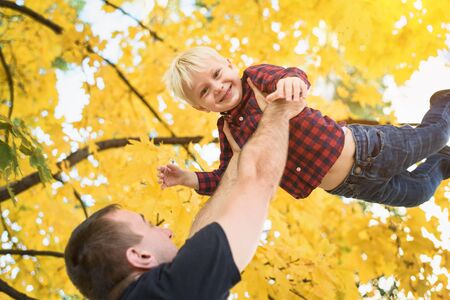 Father throws up his blond son. Yellow leaves on background. Family conceptの写真素材