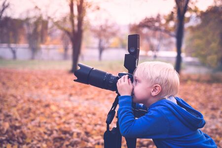 Little blond boy shoots with a large SLR camera on a tripod. Photo session in the autumn parkの写真素材