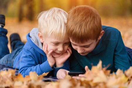 Two little brothers using smartphone, lying in yellow autumn leaves. Smiling and having fun. Fall dayの写真素材