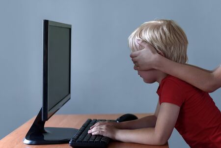 Children hands puts over eyes to a little boy who uses a computer. Dangerous content. Children and...