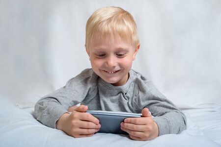 Little blond boy is watching something on a smartphone while lying in bed. Gadget Leisureの写真素材