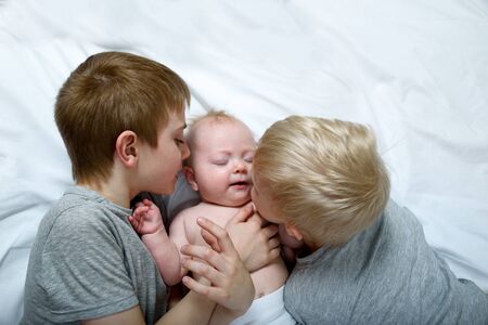 Two older brothers tenderly kiss and hug the younger child on a white bed. Happy familyの写真素材