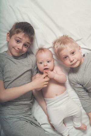 Two older brothers are lying with the youngest baby in a white bed. Happy childhood, big familyの写真素材