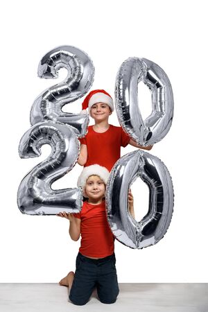 Two boys in Christmas hats are holding silver inflatable figure 2020. White background. New year conceptの写真素材