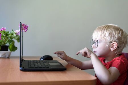 Funny blond boy in glasses sits at a laptop. Internet and preschoolerの写真素材