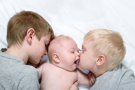 Two older brothers tenderly kiss and hug the younger child on a white bed. Happy familyの写真素材
