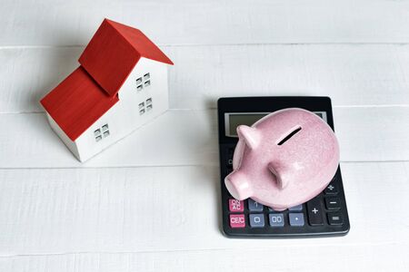 Pink pig piggy bank, calculator and breadboard model of a house with a red roof on a light background. Concept of renting, buying and selling real estate.の写真素材