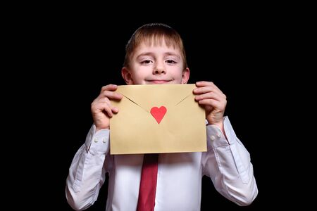 Fair-haired boy in a shirt and tie holds an envelope with a heart. Isolate on black backgroundの写真素材