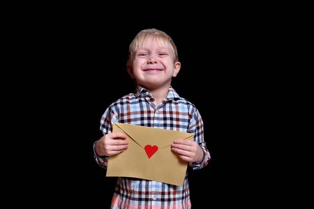 Cute blond boy holds an envelope with a red heart. Congratulations, Valentine's Day. Black backgroundの写真素材