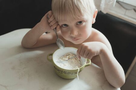 Little boy does not want to eat. Child sits at the table with a plate and spoon. Blonde kid.の写真素材