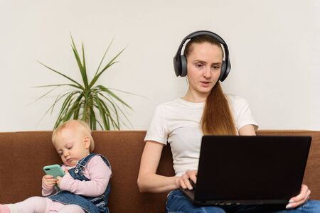 Young mother surf the Internet with laptop while small baby playing with phone. Irresponsible...