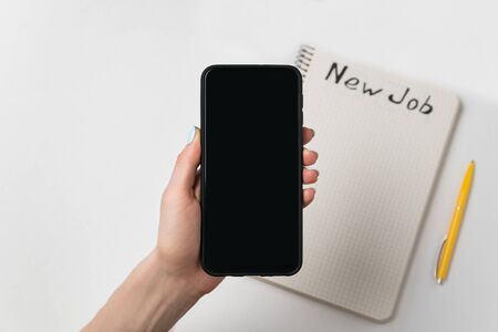 Female hand holds the phone. Pen and notebook with words New job. White background copy space.の写真素材