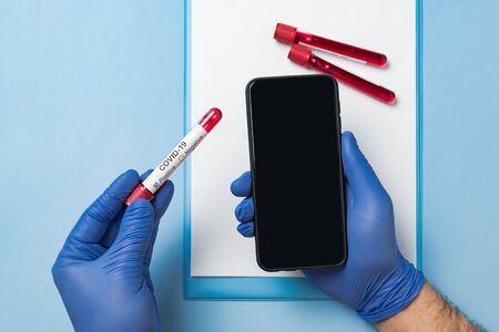 Hands in medical gloves holding smartphone with blank screen and test tube with blood. Covid-19...