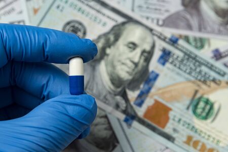 Hand in medical glove holding a pill on background of money. Cost of medicines and treatment.の写真素材