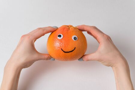 Grapefruit with funny face in female hands on white background. Orange with Googly eyes and painted smile.の写真素材