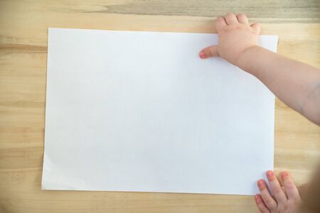 Childrens hand holding blank white paper sheet on wooden background. Copy space. Template. Mockup.