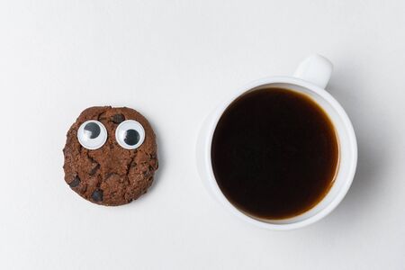 Cup of tea and chocolate chip cookie with funny face and Googly eyes. Coffee mug on white background...