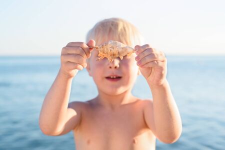 Cute blond boy holding a shell on sea background. Holiday of sea with son.の写真素材