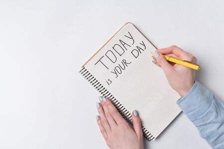 Motivational inscription TODAY IS YOUR DAY. Top view of woman writing in Notepad over white...