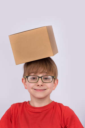 Boy in red shirt and glasses smiling and holding cardboard box on his head. Vertical frame.の写真素材