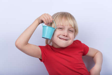 Boy with sly face holding small bucket. Blond child in red shirt on white background.