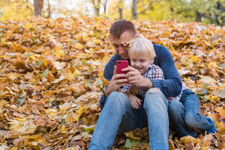 Dad teaches child to take pictures on phone. Father and son sitting in autumn Park